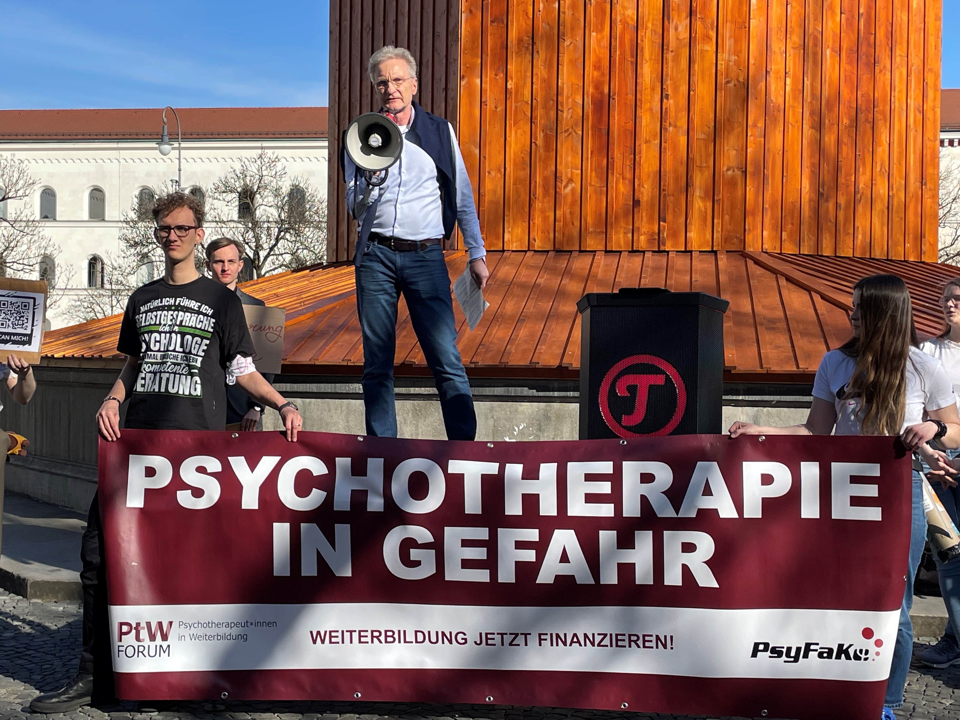 Ein Mann steht bei der Demonstration auf einem Podest und hält eine Rede mit einem Megafon. Davor halten Studierende ein großes Demo-Banner.